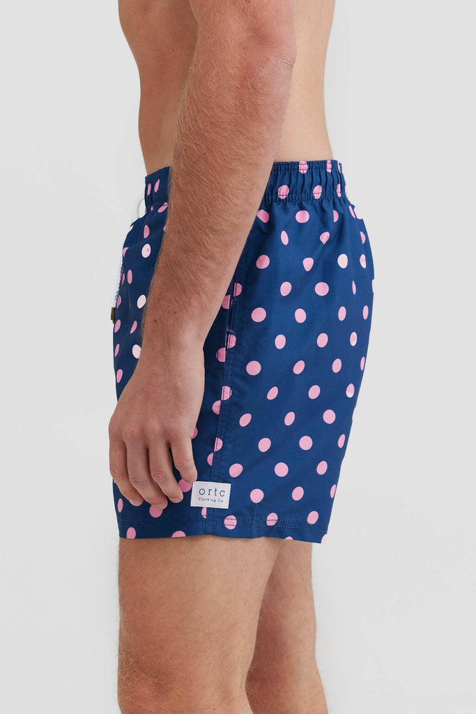 Side view of navy boardshorts with pink polka dots on them and white ortc logo