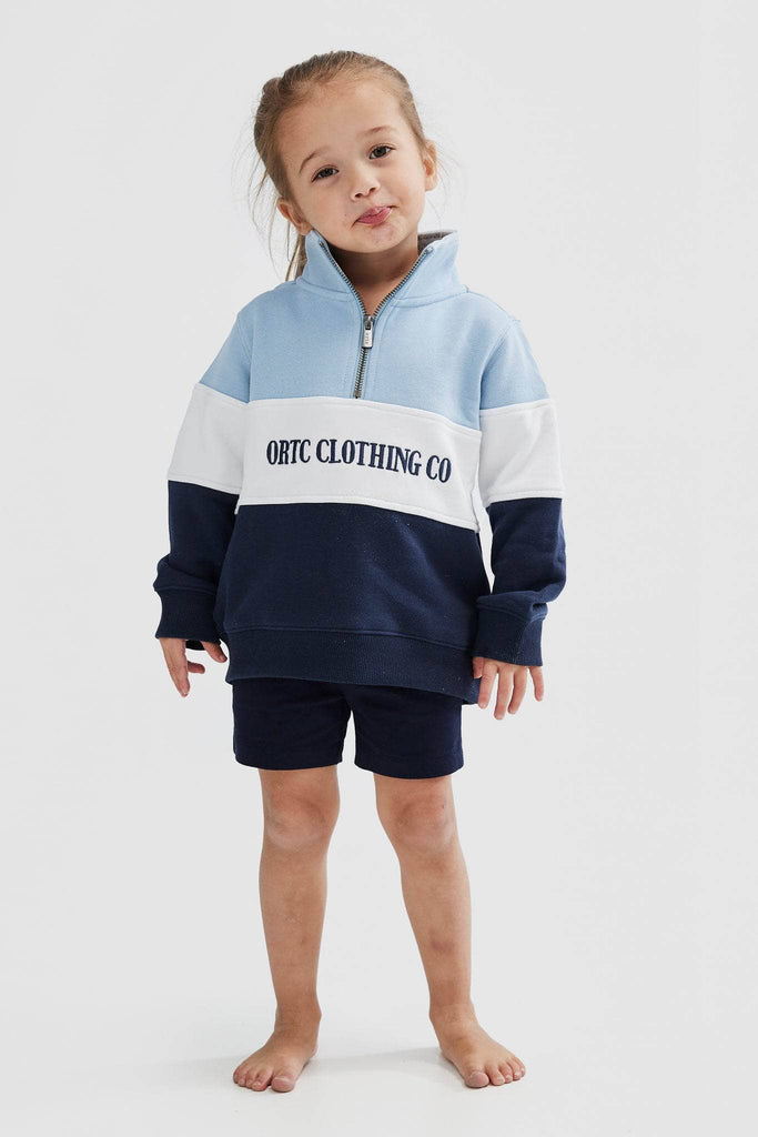 Child model wearing quarter zip jumper that has a light blue top panel, white middle panel with navy embroidered ortc Clothing Co and navy base panel