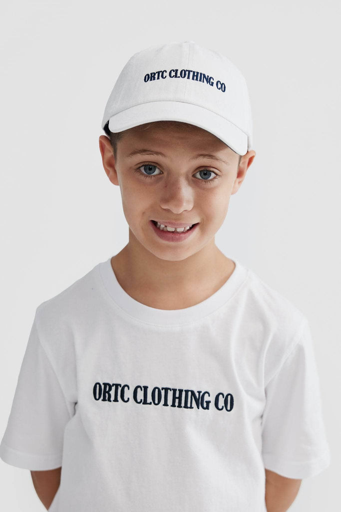 Child model wearing white baseball cap with navy embroidered ortc clothing co